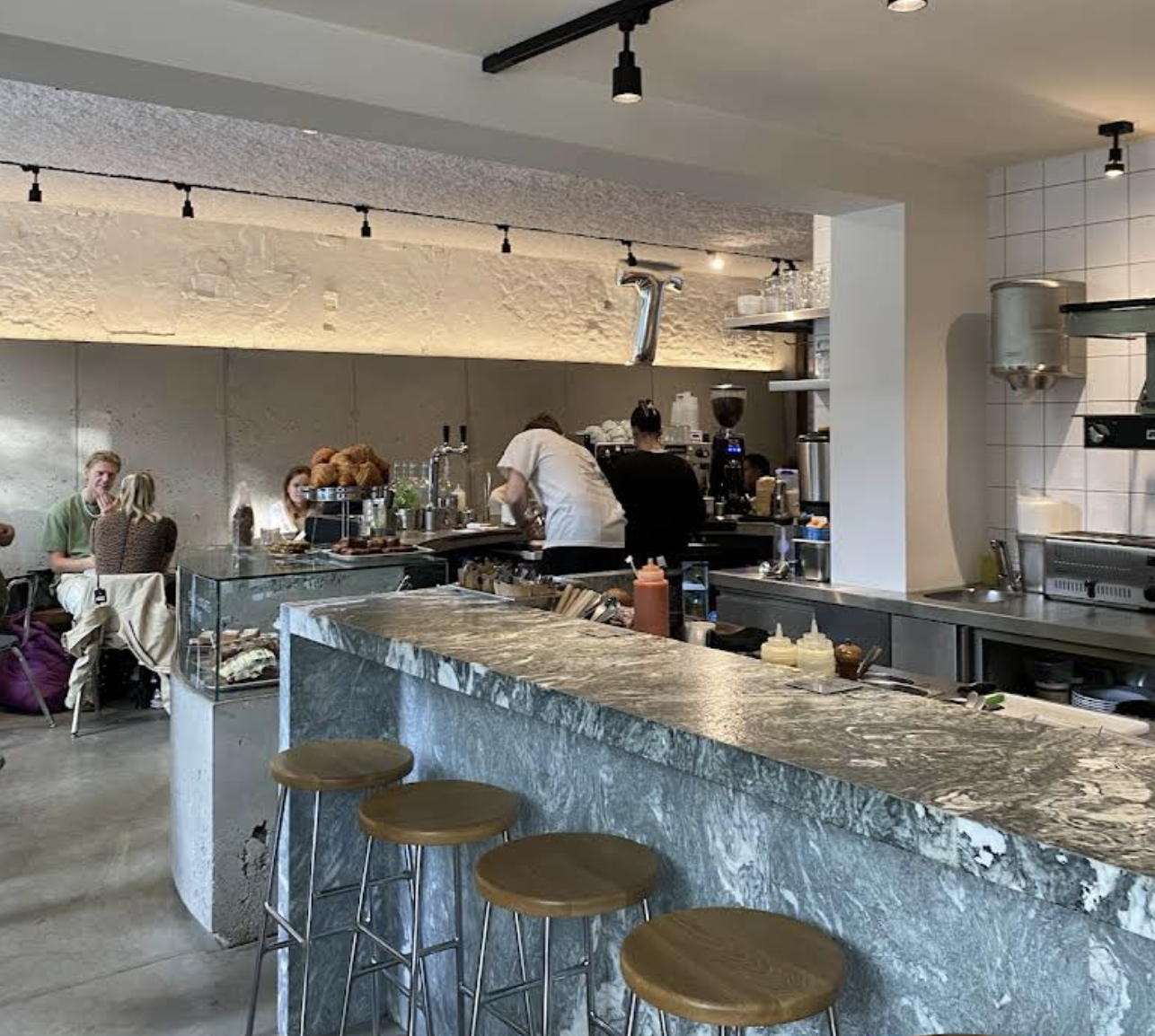 Inside a modern coffee shop with a marble counter, bar stools, and people seated at tables. The counter has squeeze bottles, and behind it, baristas prepare drinks with coffee equipment. The decor features textured ceiling and wall, with pendant and 