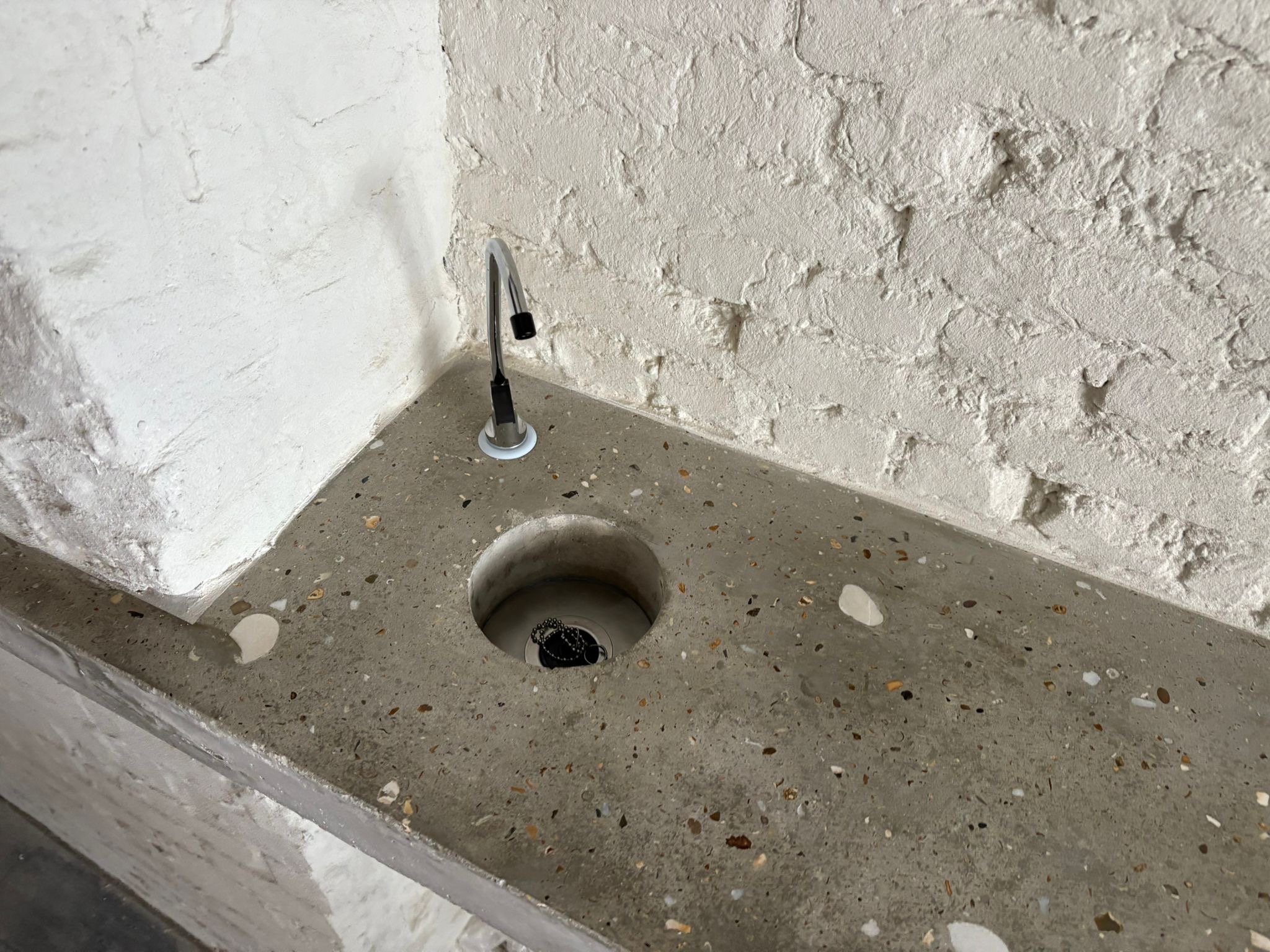 Close-up view of a rough textured wall in a corner with a metallic plumbing pipe and a black stopper, set above a concrete surface with small debris.