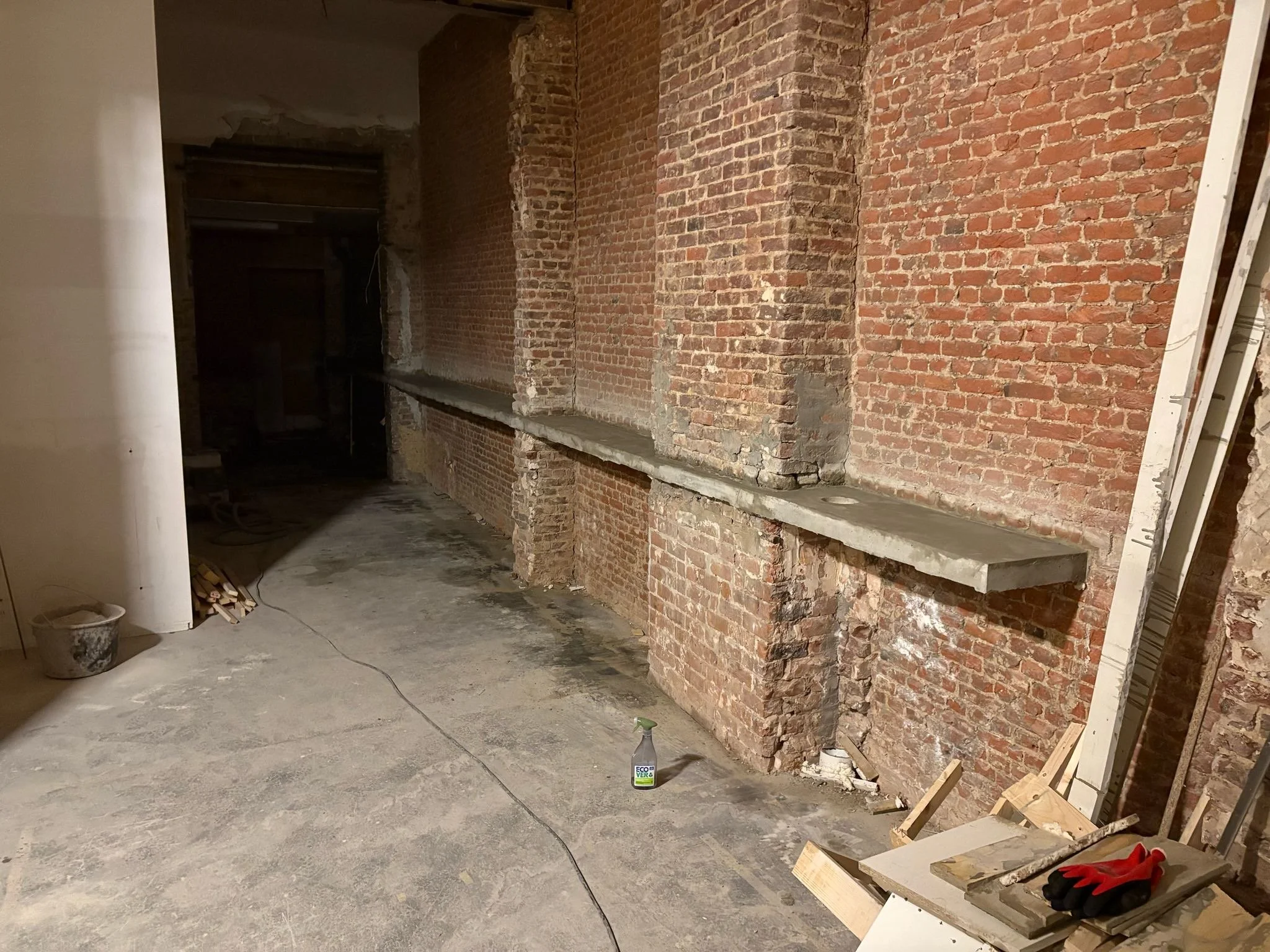 An unfinished interior space with exposed brick walls, concrete floor, bookshelves, and construction tools.