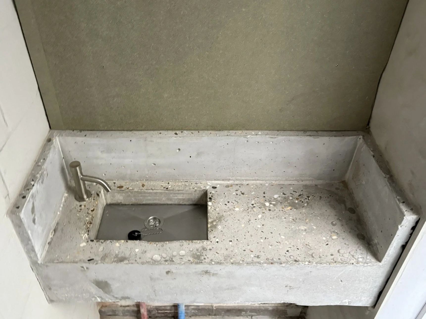 Construction area with a small stainless steel sink with a chain and plug, set into a speckled gray concrete countertop. The surrounding area shows unfinished surfaces with visible dust, debris, and unpainted or unfinishe drywall walls.