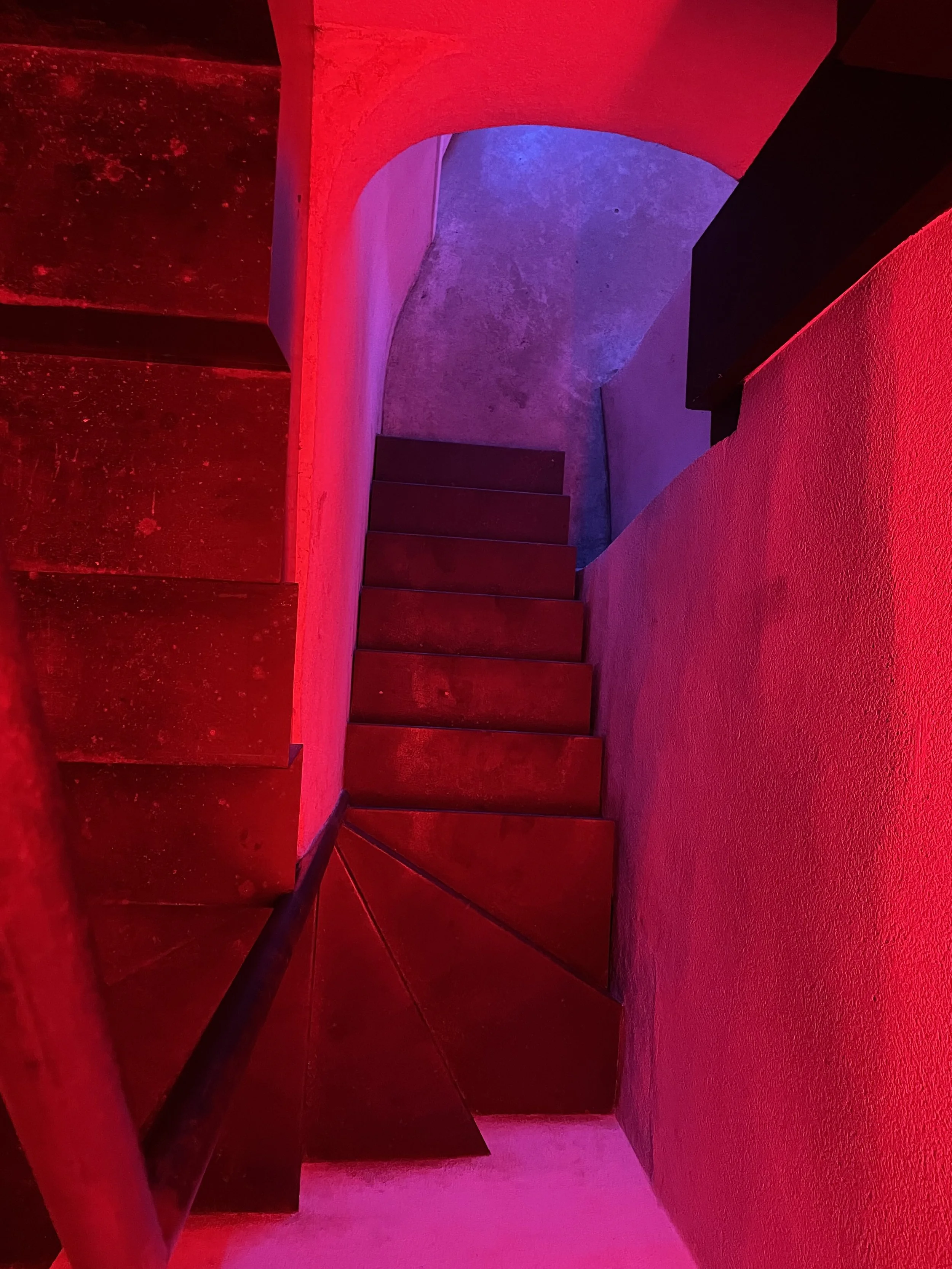 A staircase with wooden steps and handrails, lit with red and purple accent lighting, leading upward through a modern, colorfully lit space.
