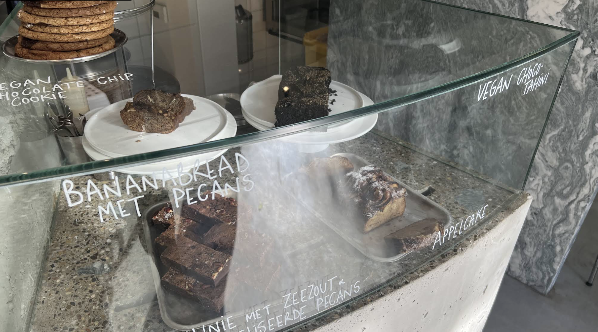 Glass display case containing chocolate desserts with white handwritten labels: 'Vegan Chocolate Chip Cookie', 'Vegan Choco-Tahini', 'Banana Bread with Pecan', and 'Apple Cake'.