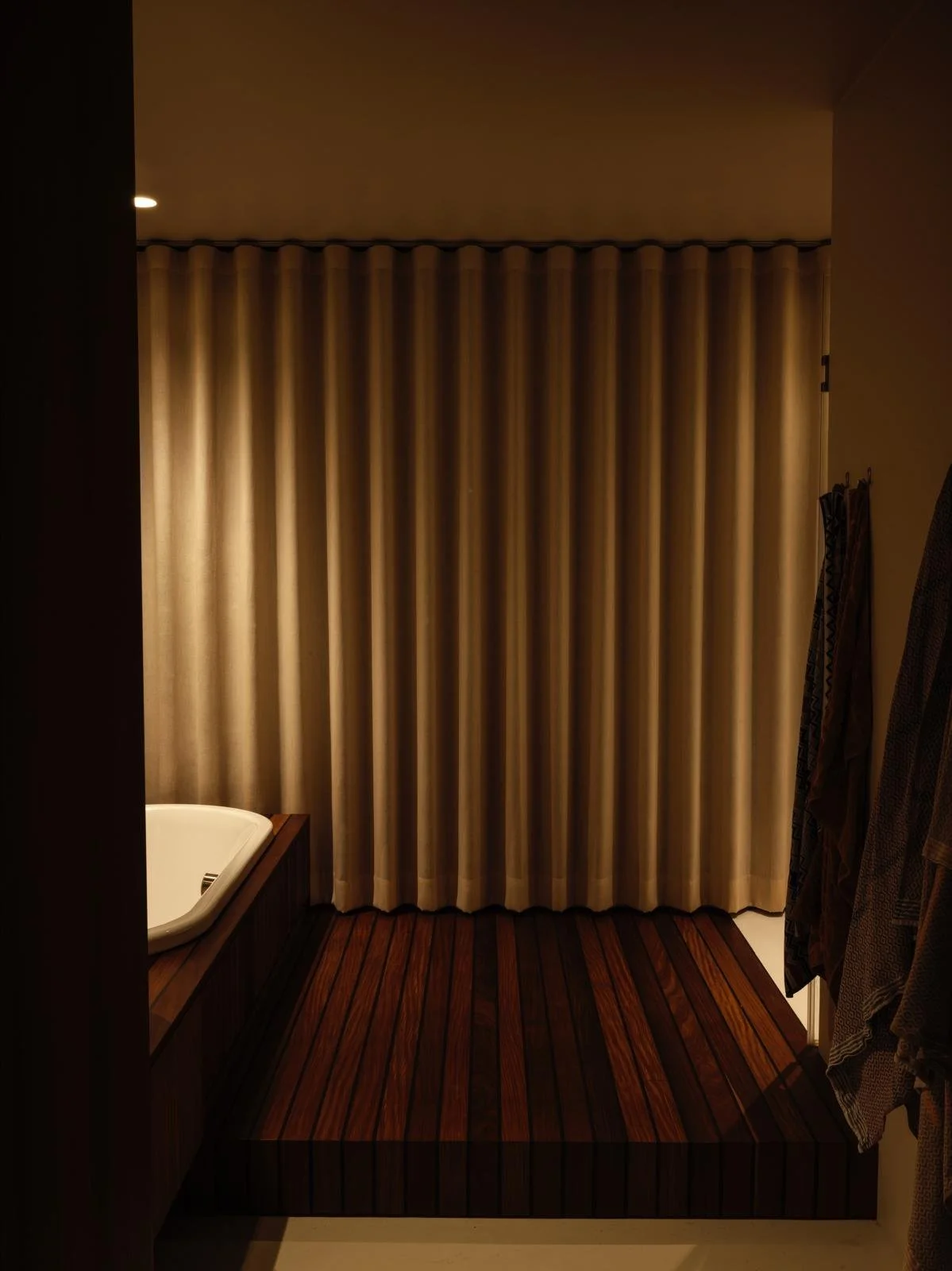 Interior of a bathroom with a corner bathtub, beige curtains, wooden flooring, and hanging towels.