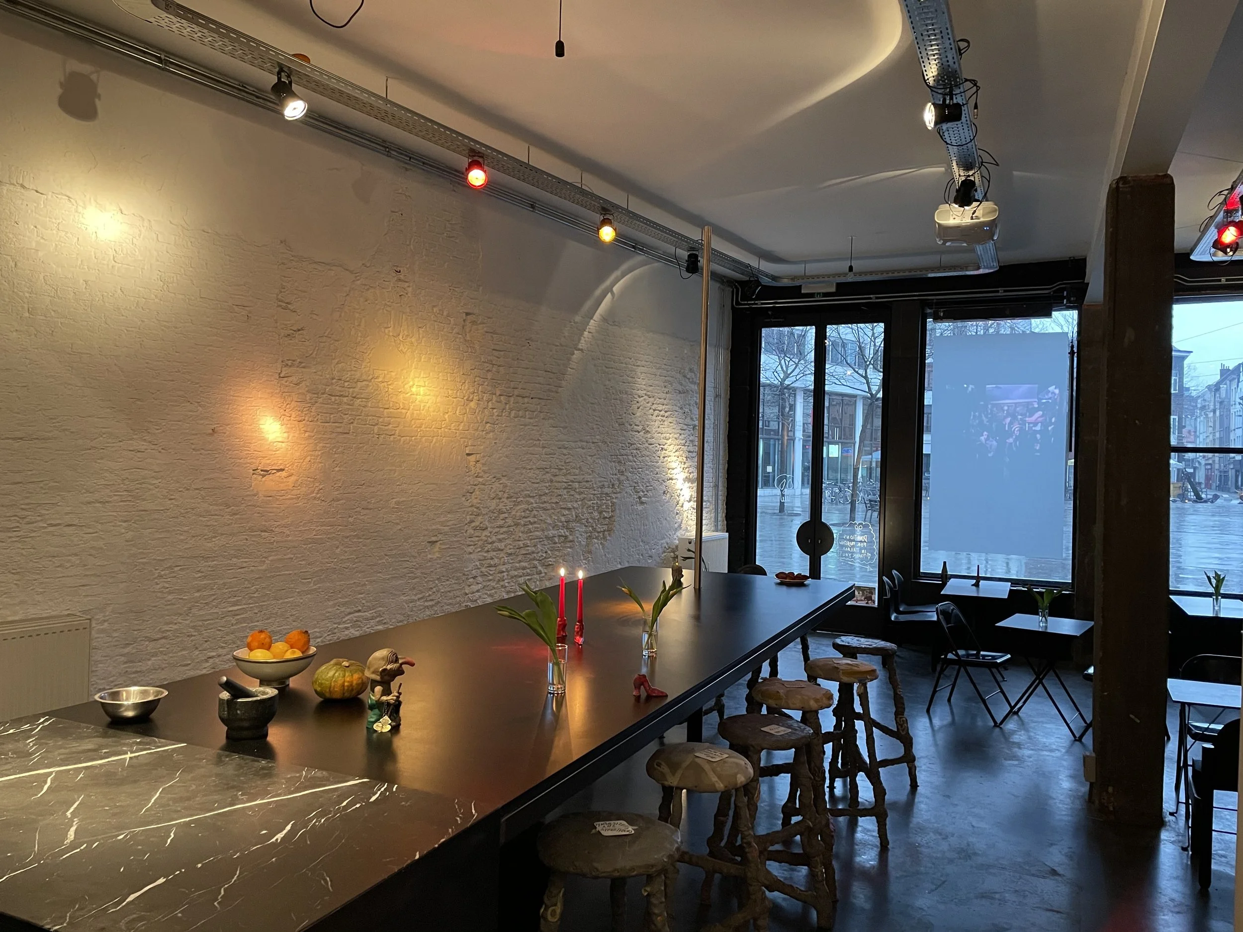 Interior of a modern space with a black marble-topped table, small bowls, some decorative figurines, and candles. Light from large windows illuminates the room, which has white brick walls, a ceiling with track lighting, and a row of rustic stools al