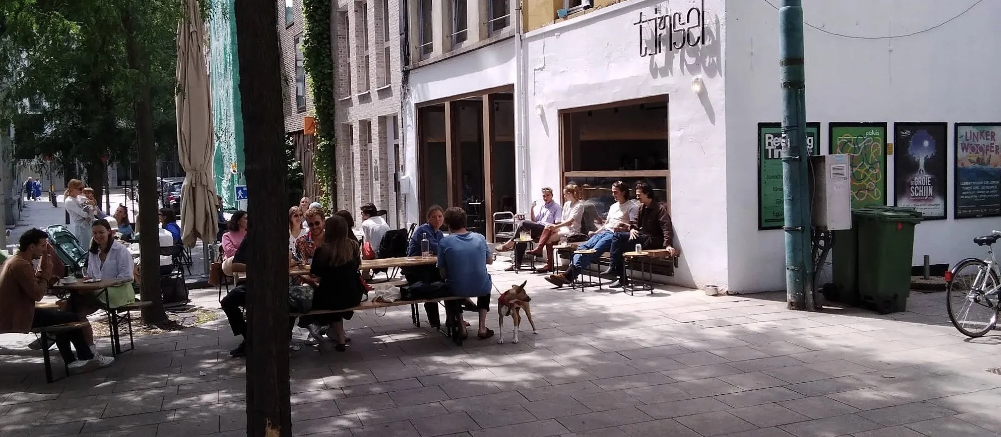 People sitting and chatting outside a cafe on a city sidewalk during the daytime, with a dog on a leash nearby, trees, and buildings in the background.