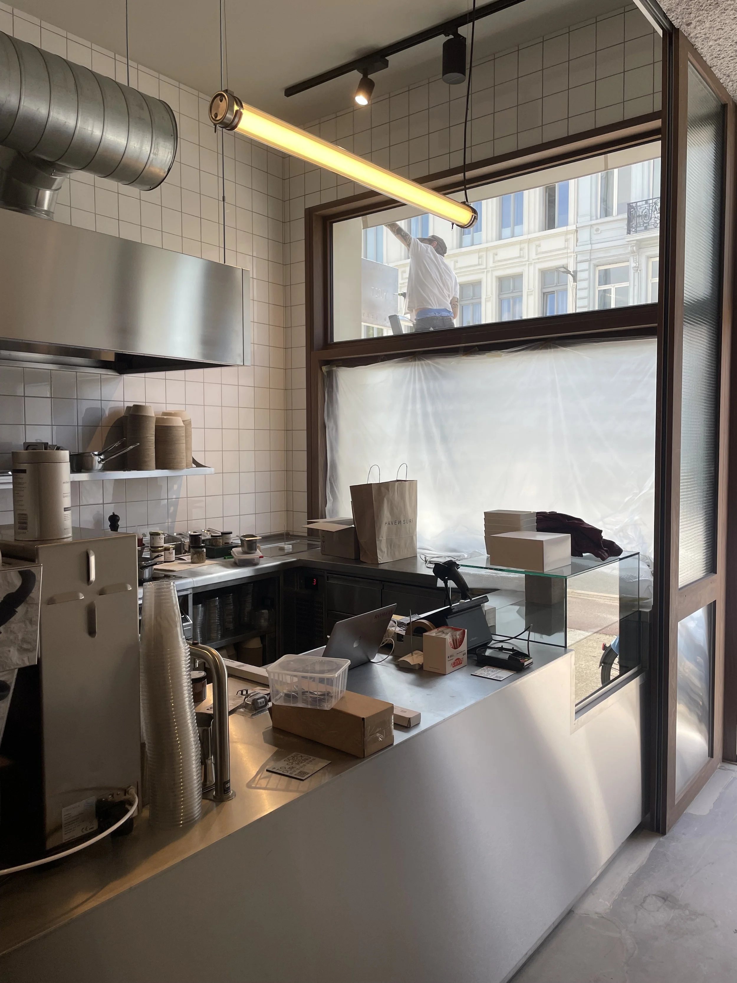 Interior of a modern coffee shop with white tiled walls, a counter with various cups and supplies, and a large window revealing a person cleaning outside the building.