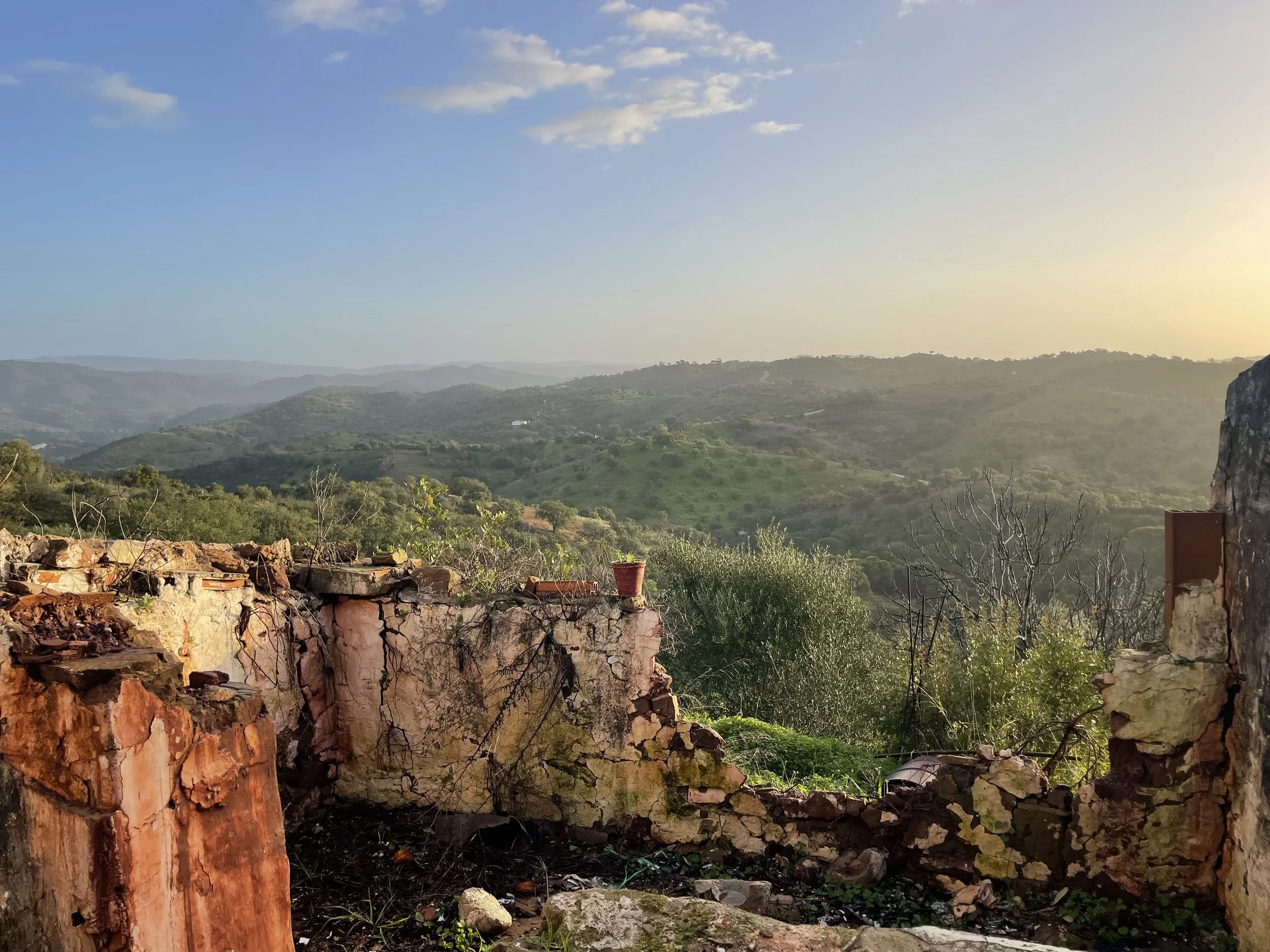 View of green hills and mountains under a blue sky with scattered clouds, seen from a rustic stone and brick wall in the foreground.
