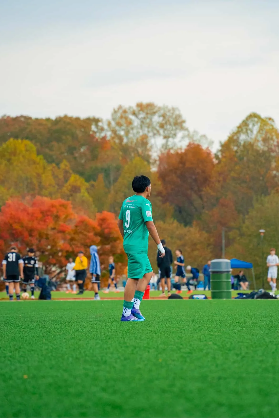 A young man in a green sports uniform stands on a lush green artificial soccer field, with autumn trees in the background. Several other players and individuals are visible in the distance, some wearing dark jerseys, engaged in activities or watching. The scene captures a sports practice or game in progress during fall.