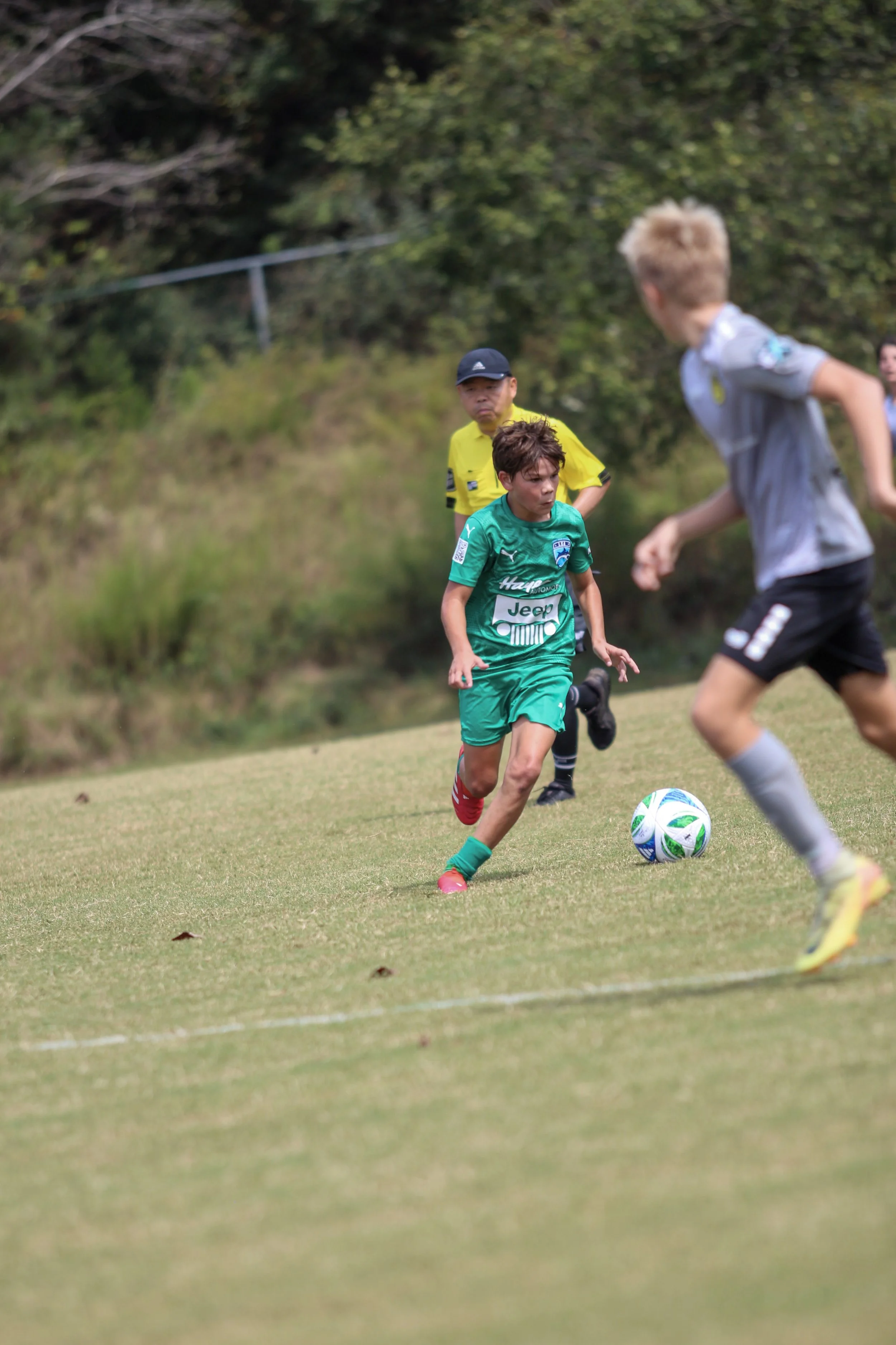 A young boy in a green sports uniform playing soccer on a grassy field, with a soccer ball at his feet and other players and a referee in the background.