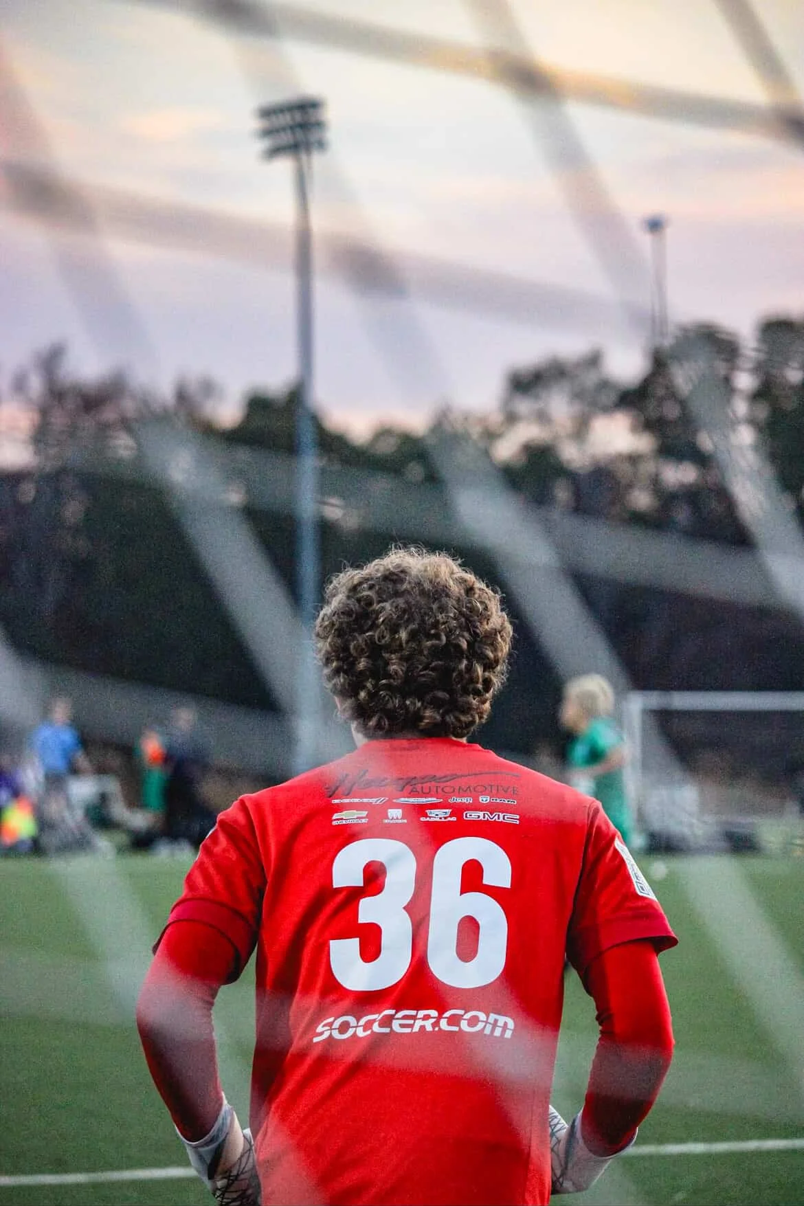 A soccer player with curly hair, wearing a red jersey with the number 36 and the logo soccer.com on the back, standing on a field and looking towards the goal during a game at dusk.