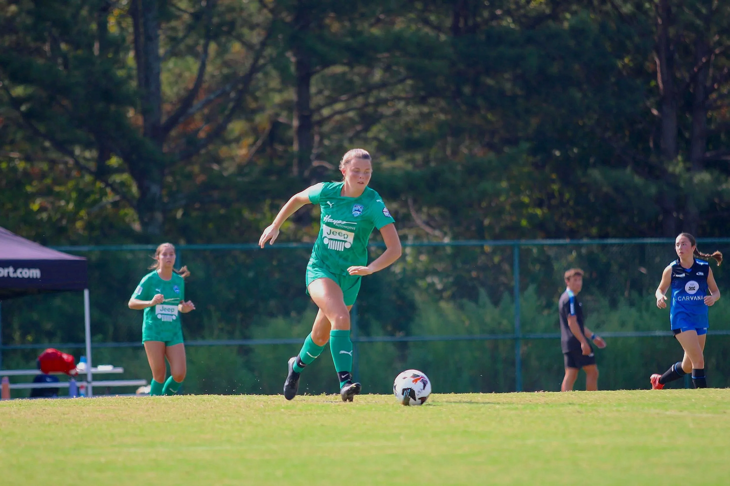A woman playing soccer on a grassy field during daytime, wearing a green uniform with logos, with other players and trees in the background.