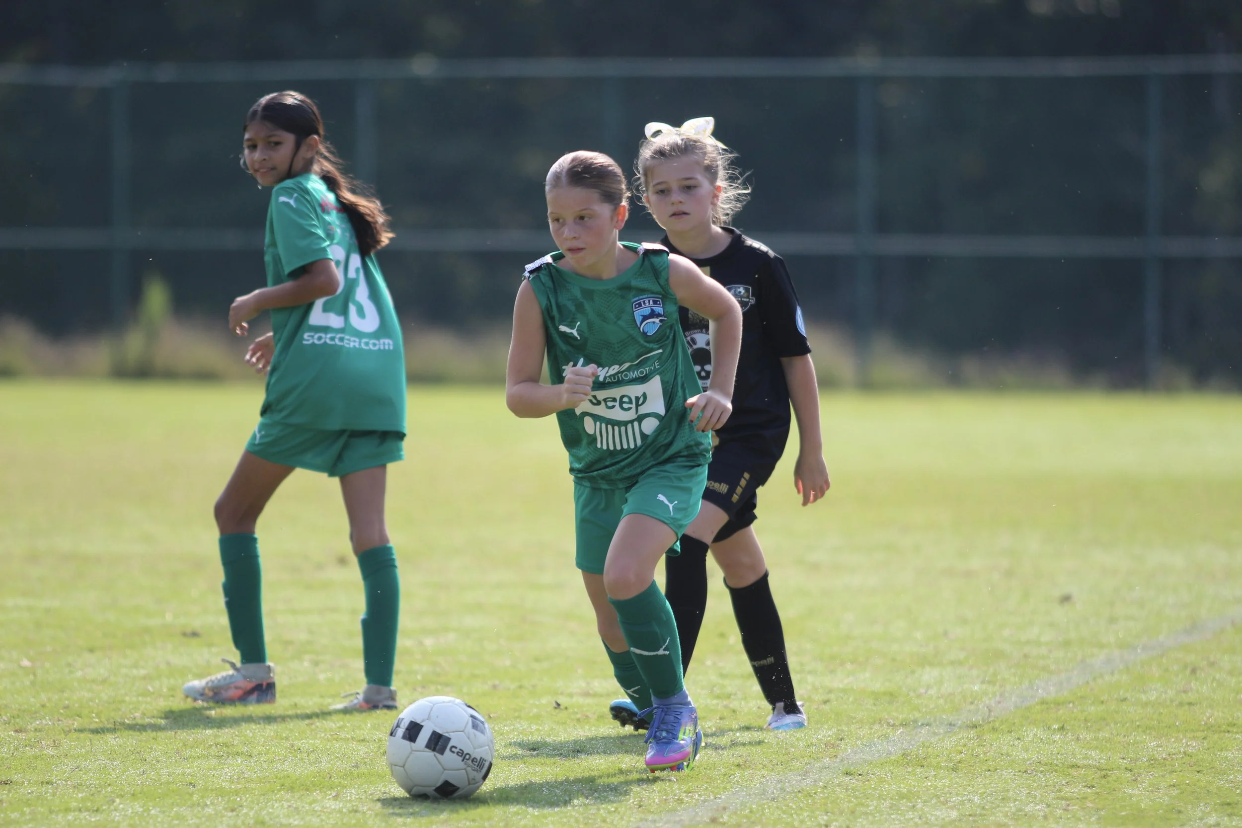 Three young girls in sports uniforms playing soccer on a field, with one girl running towards a soccer ball in the foreground.