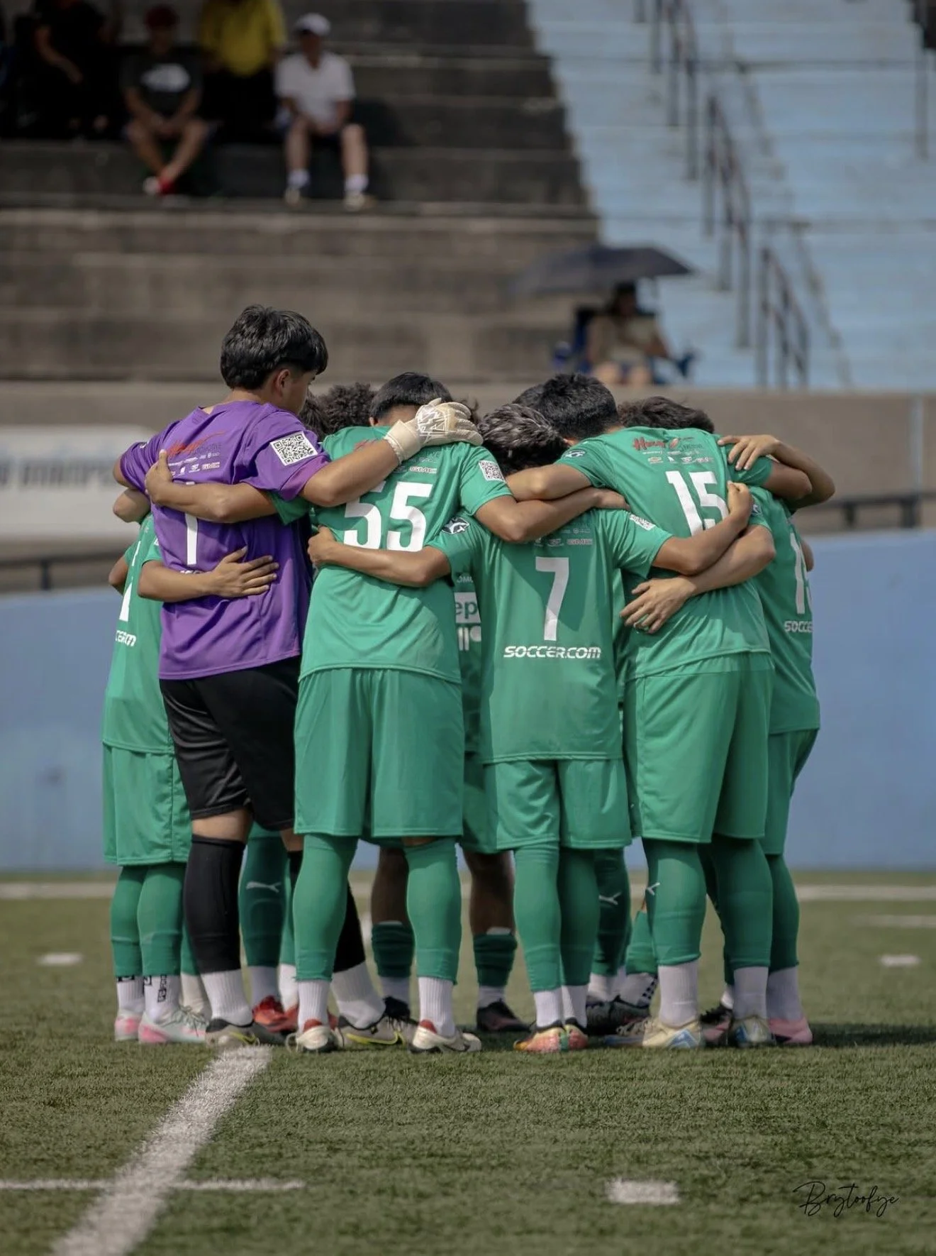 A group of soccer players in green uniforms huddled together on the field, with a goalkeeper in purple joining their huddle.