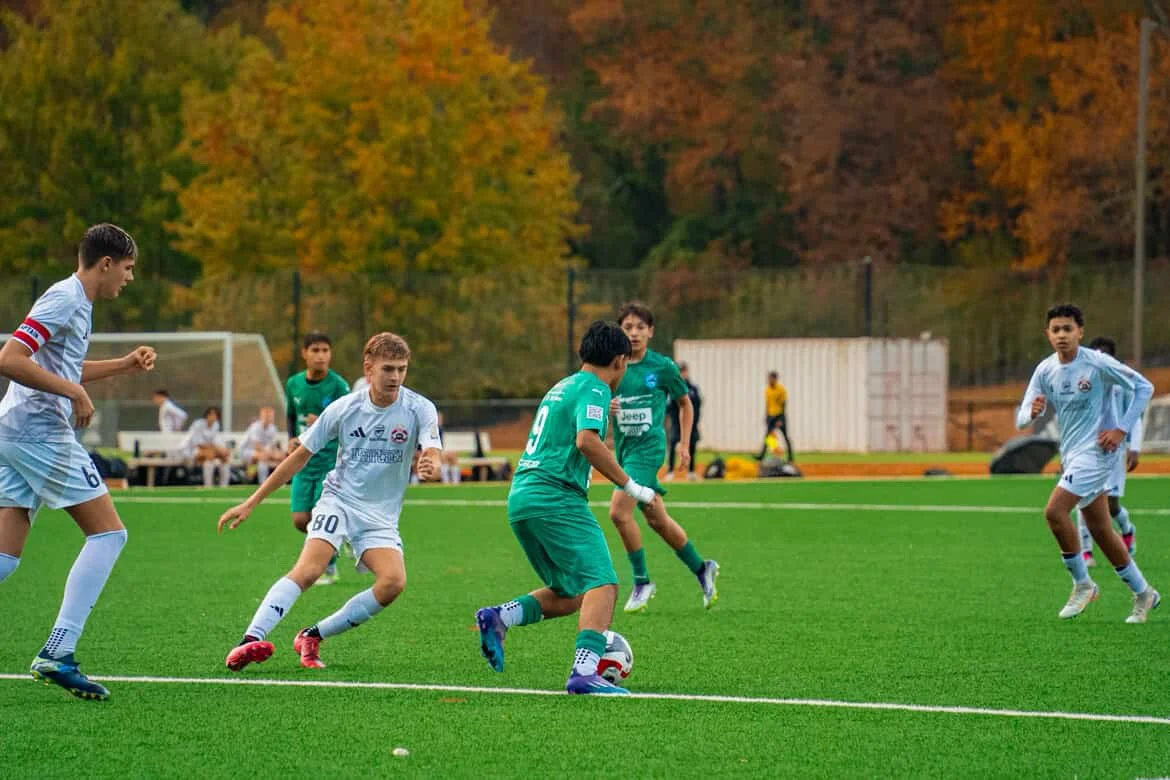 Young boys playing soccer on a field with autumn trees in the background.