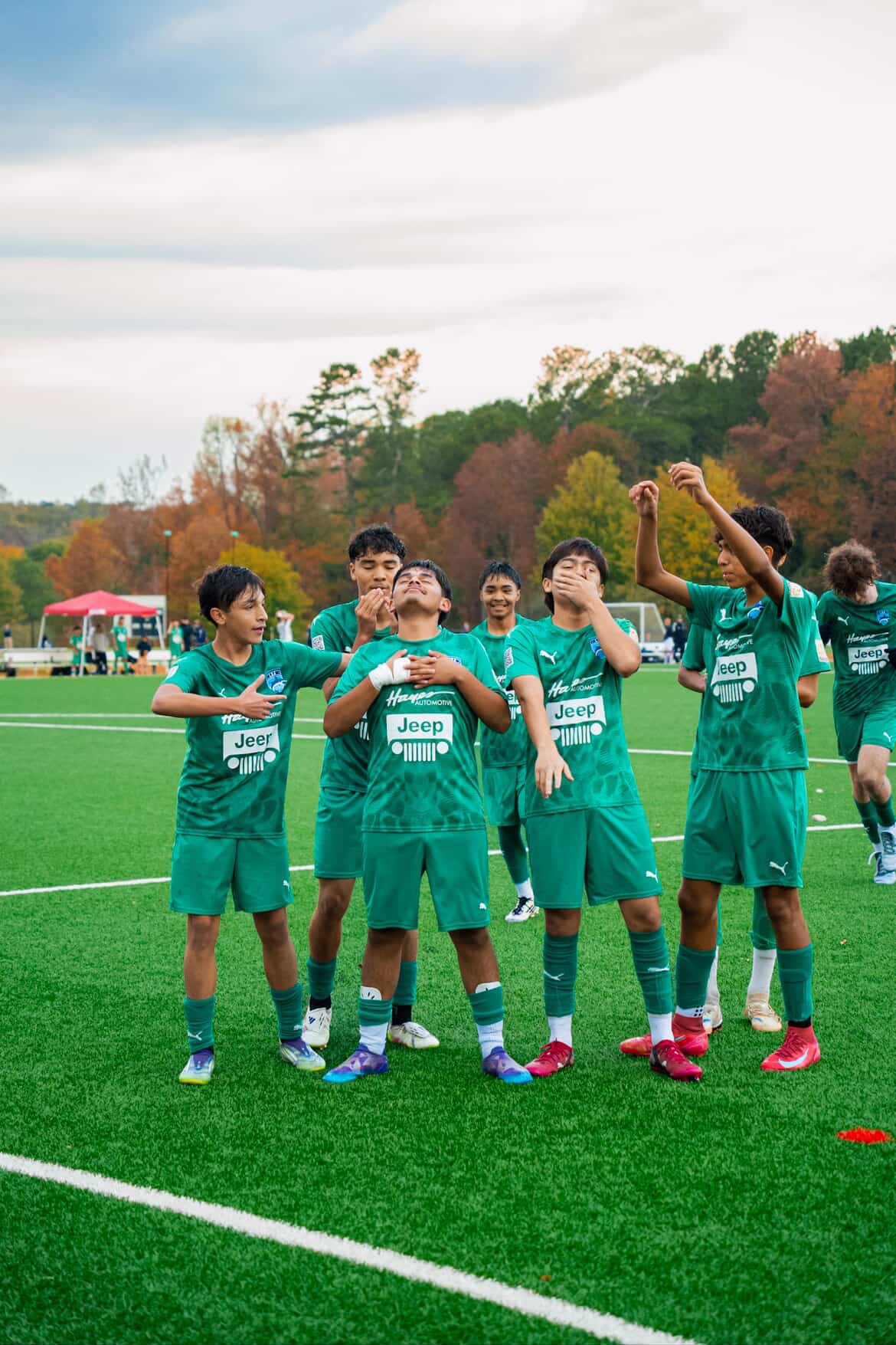 Group of young male soccer players in green jerseys on a soccer field, some showing expressions of joy and celebration, with trees with fall foliage and cloudy sky in the background.