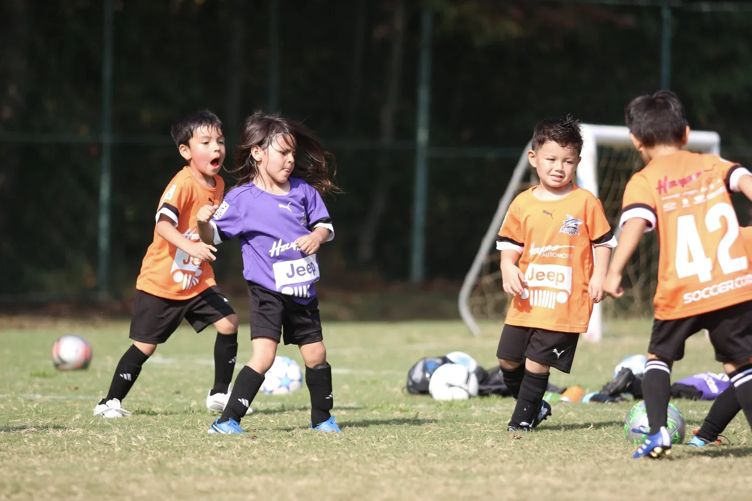 Young children playing soccer on a field, wearing colorful jerseys, with some children looking focused and others appearing surprised, and soccer balls scattered on the grass.