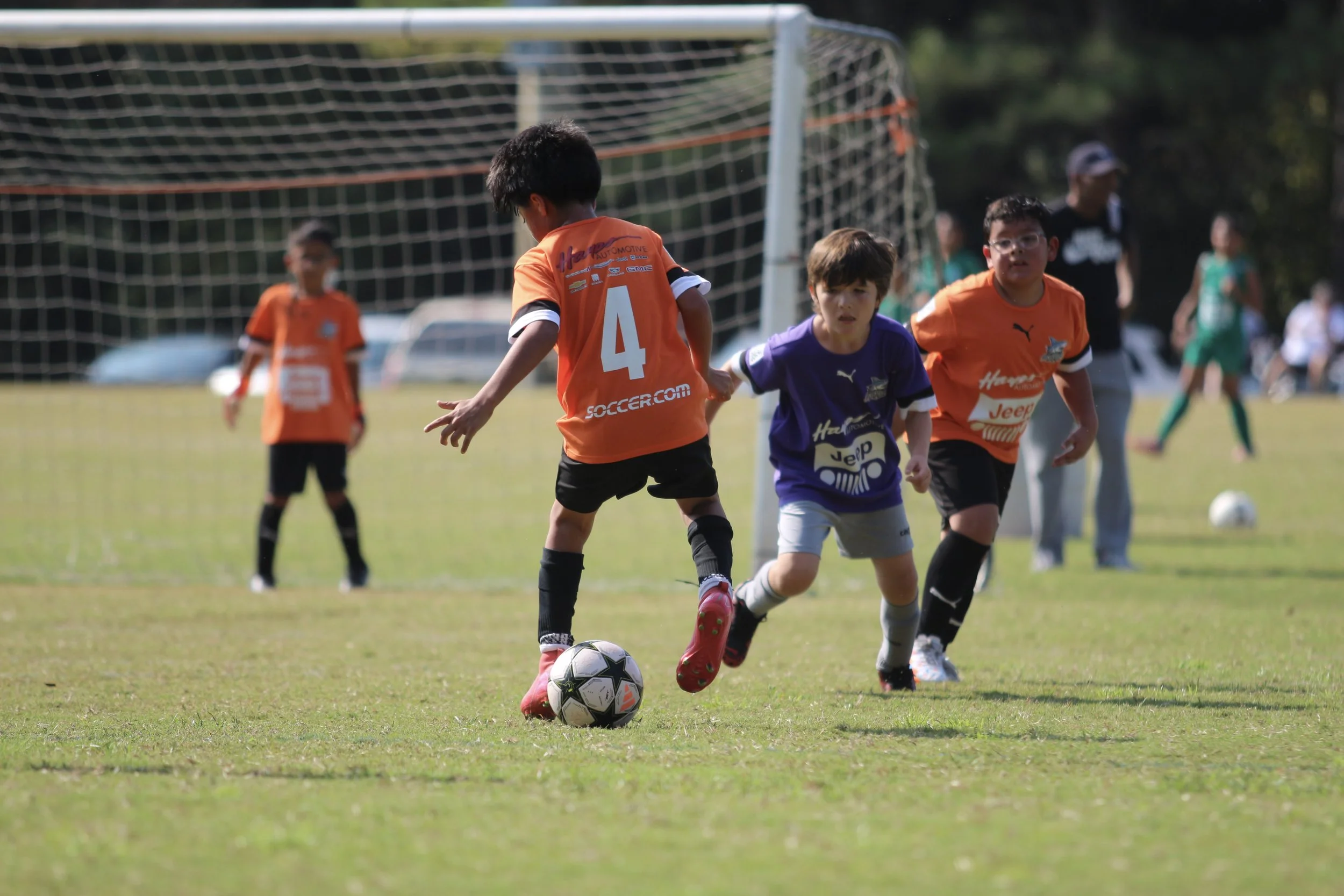 Children playing youth soccer on a field, with a soccer ball near two kids in the foreground and goalpost in the background.
