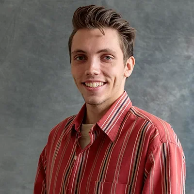 A young man with light skin and brown hair styled to the side, smiling and wearing a red and beige striped button-up shirt, standing in front of a gray textured background.