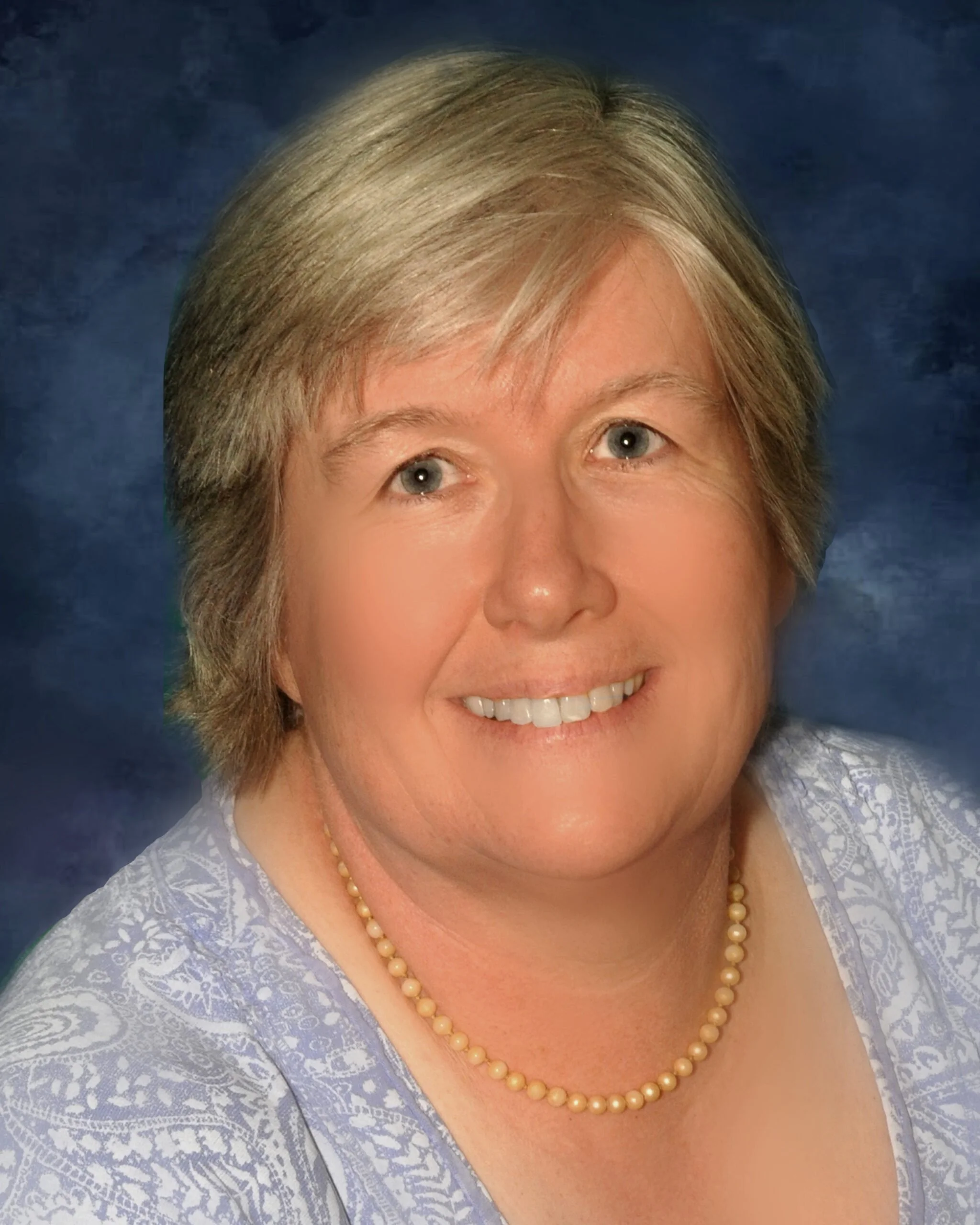Close-up of a smiling woman with blonde hair, wearing a pearl necklace and a light-colored lace top, against a dark blue background.