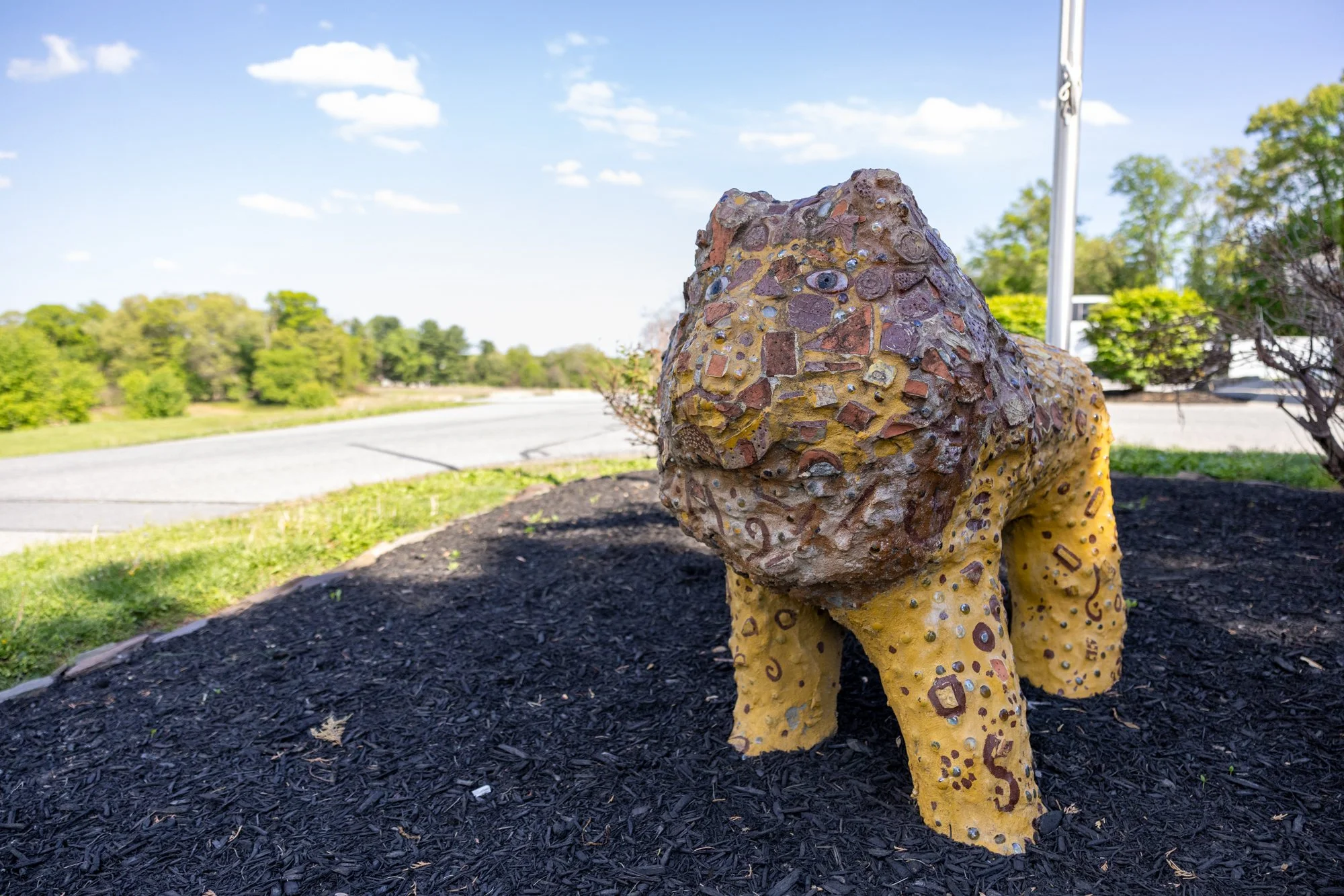 A sculpture of a humanoid figure made of multicolored stones and embedded with beads, sitting outdoors on black mulch with green trees and a parking lot in the background.