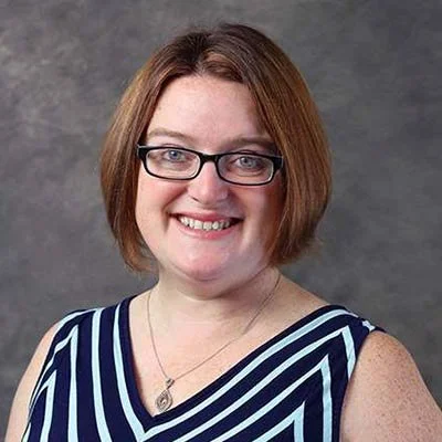A woman with shoulder-length brown hair wearing glasses and a blue and white striped sleeveless top, smiling against a gray background.