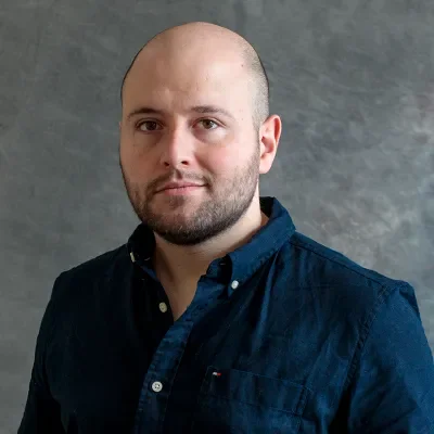 Portrait of a man with a bald head, beard, and wearing a dark button-up shirt, standing against a gray background.