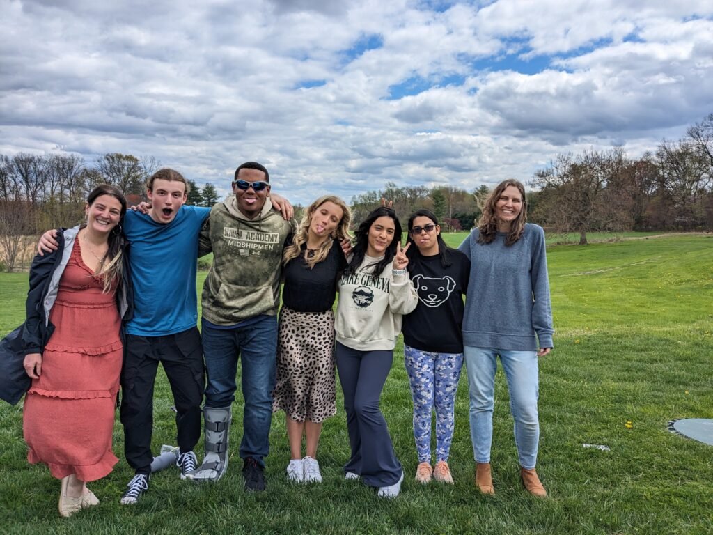 Group of seven young adults standing outdoors on a grassy field, smiling and posing for the photo with trees and a cloudy sky in the background.
