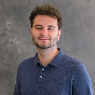 A young man with dark hair and a beard, wearing a blue polo shirt, smiling at the camera against a gray background.