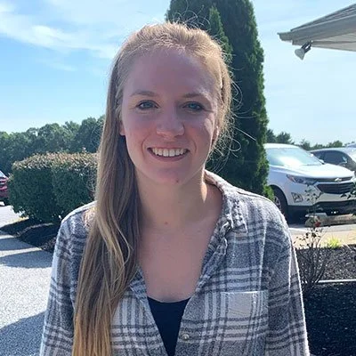 Young woman with long blonde hair smiling outdoors on a sunny day, wearing a gray plaid shirt, in front of greenery and parked cars.