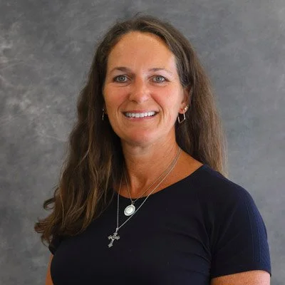 A woman with long, wavy brown hair, smiling, wearing a black top, silver earrings, a necklace with a round pendant, and a cross necklace, standing against a gray background.