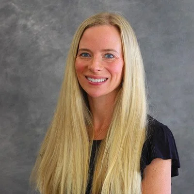 A young woman with long blonde hair, smiling, wearing a black top, standing against a gray background.