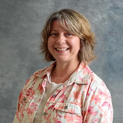 A woman with short, wavy blonde hair, smiling, wearing a light-colored shirt with a pink floral pattern, standing against a gray textured background.