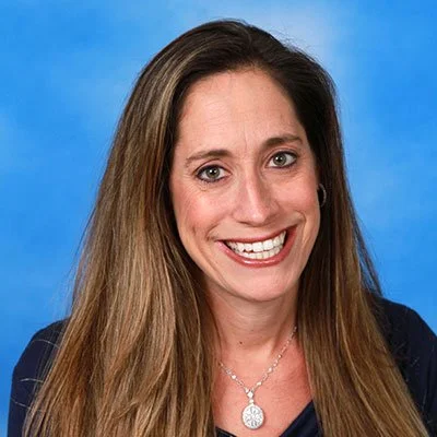 Portrait of a woman with long brown hair, smiling, wearing a necklace with a round pendant, against a blue background.
