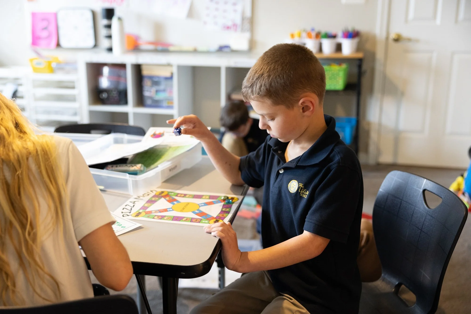 A young boy playing a board game called 'Pig Game' at a table in a classroom. He is holding a die and focused on his turn. The classroom has shelves with various supplies and colorful posters on the wall.