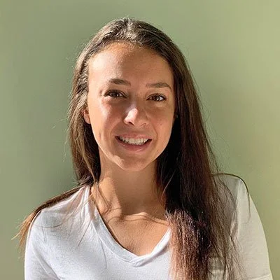 A young woman with long brown hair smiling at the camera, wearing a white top against a light green background.