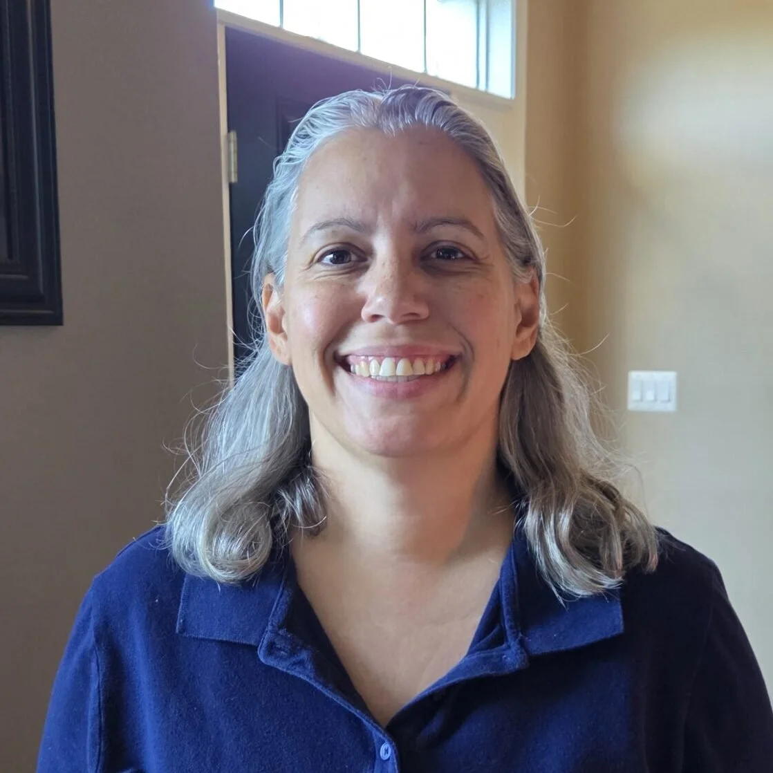 A smiling woman with shoulder-length gray hair, wearing a blue collared shirt, in a well-lit room with a window and yellow walls.