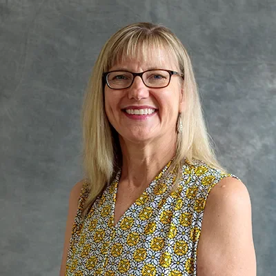 A woman with blonde hair, glasses, and a patterned sleeveless blouse smiling at the camera.
