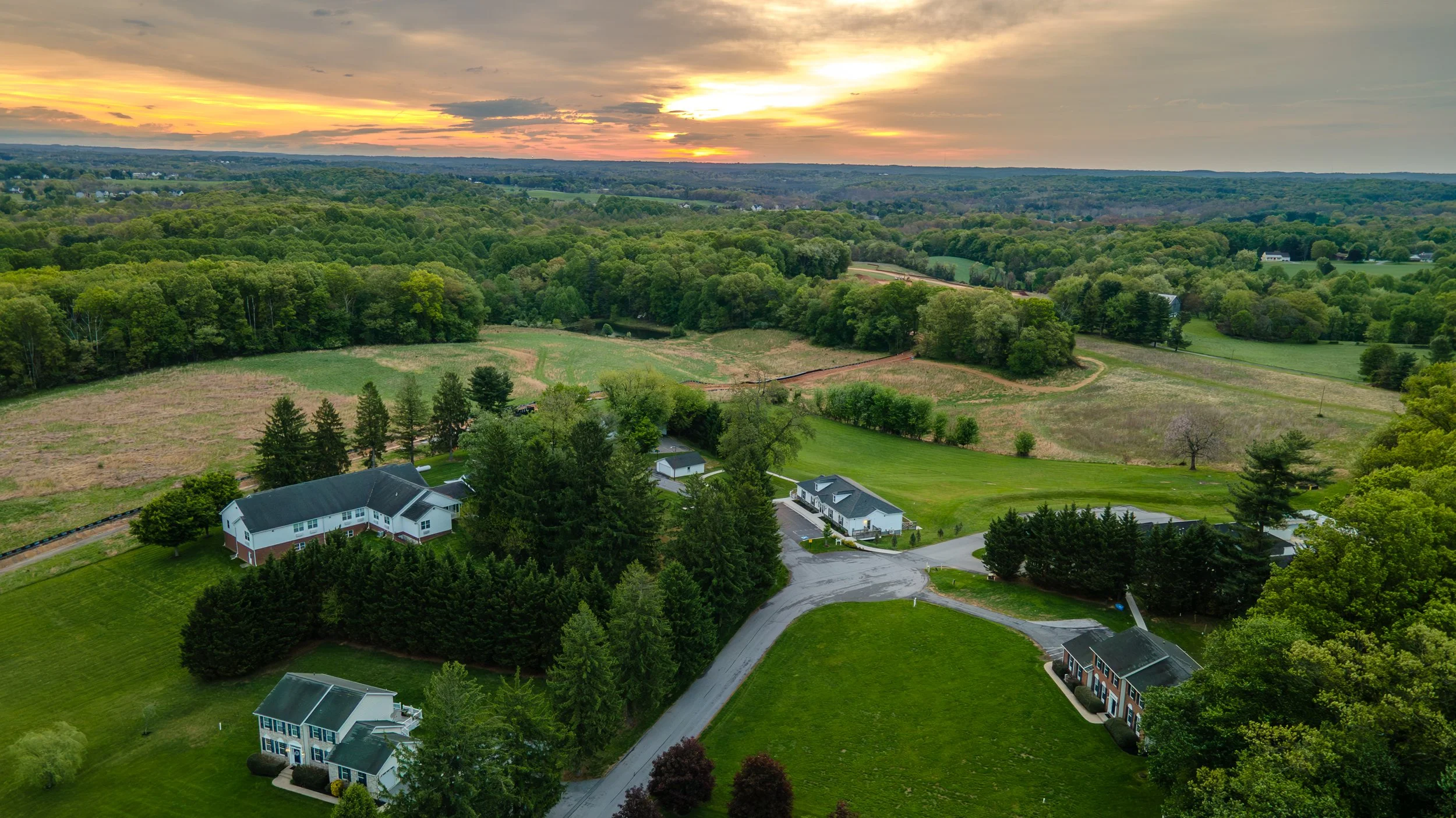 Aerial view of a rural area at sunset with houses, trees, green fields, and a winding driveway.