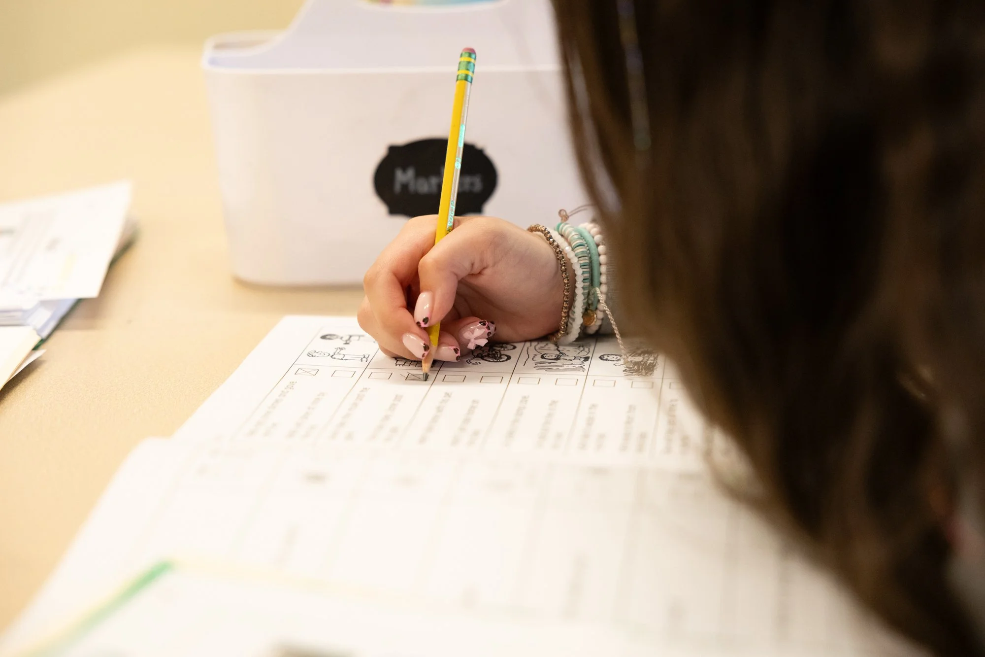 A person with manicured nails and multiple bracelets, using a yellow pencil with colorful bands to mark answers on a printed questionnaire or worksheet, at a table with a white box in the background.