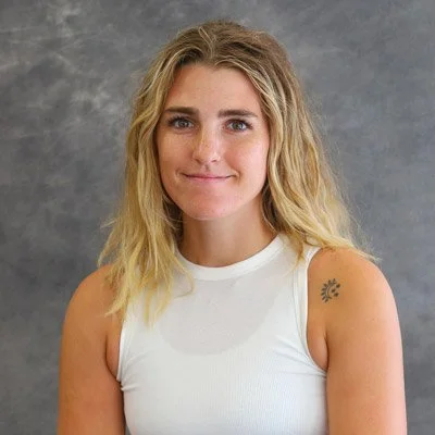 A young woman with wavy blonde hair, wearing a sleeveless white top, and a small tattoo on her left shoulder, standing in front of a gray backdrop.