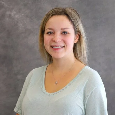 A young woman with blonde hair smiling at the camera, wearing a light-colored shirt, against a gray background.