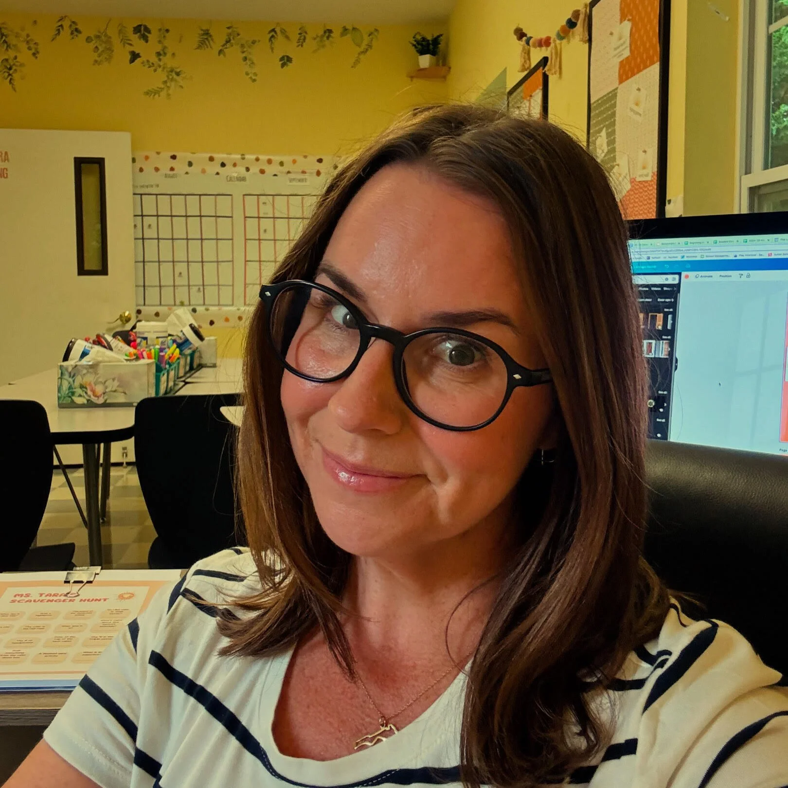 A woman with glasses and brown hair smiling at the camera in a classroom or office setting.