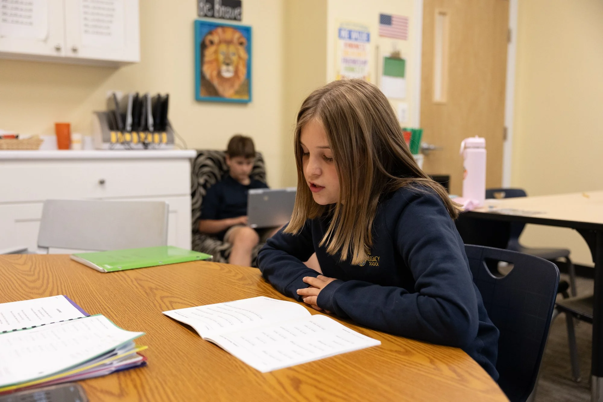 A young girl studying at a table with open notebooks and papers in front of her, while a young boy is sitting in the background using a tablet in a classroom with educational posters and decorations on the walls.
