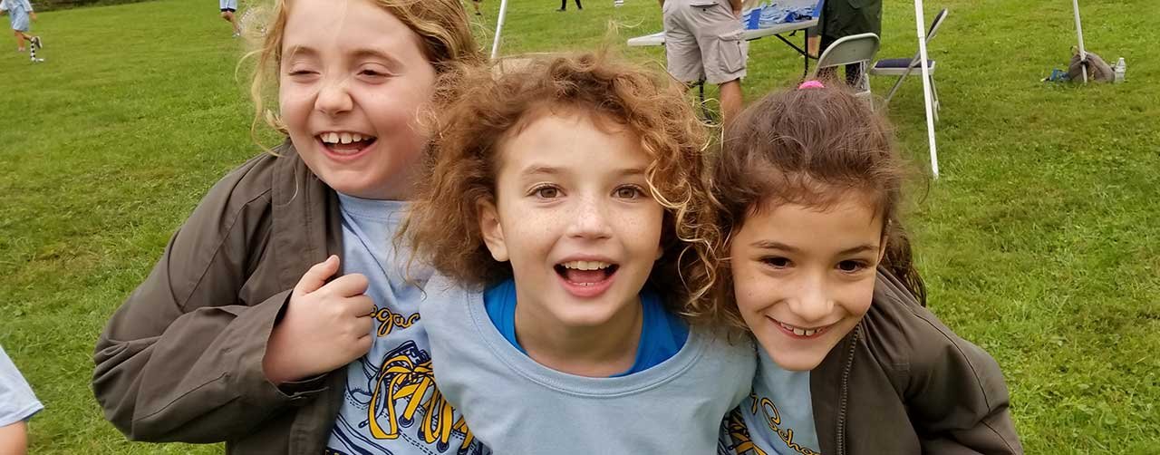 Three young girls smiling and posing closely together outdoors on a grassy field, with tents and people in the background at an event or gathering.