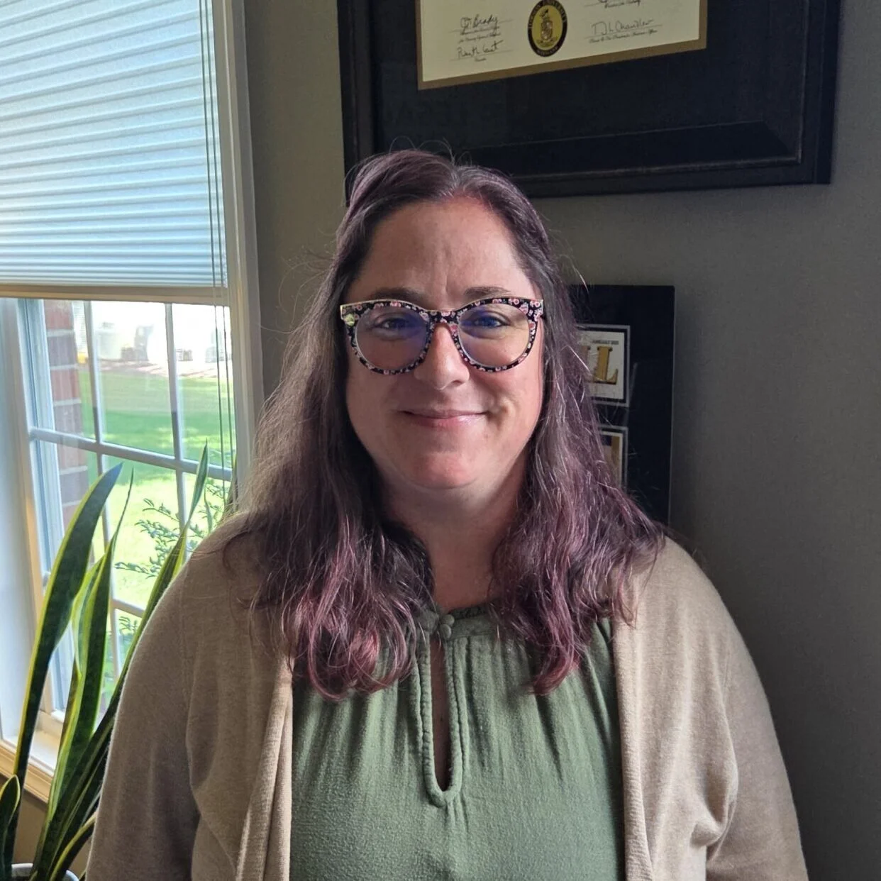 A woman with long wavy purple hair wearing glasses with a floral pattern, a green top, and a beige cardigan, standing indoors near a window with blinds, framed awards or certificates on the wall behind her.