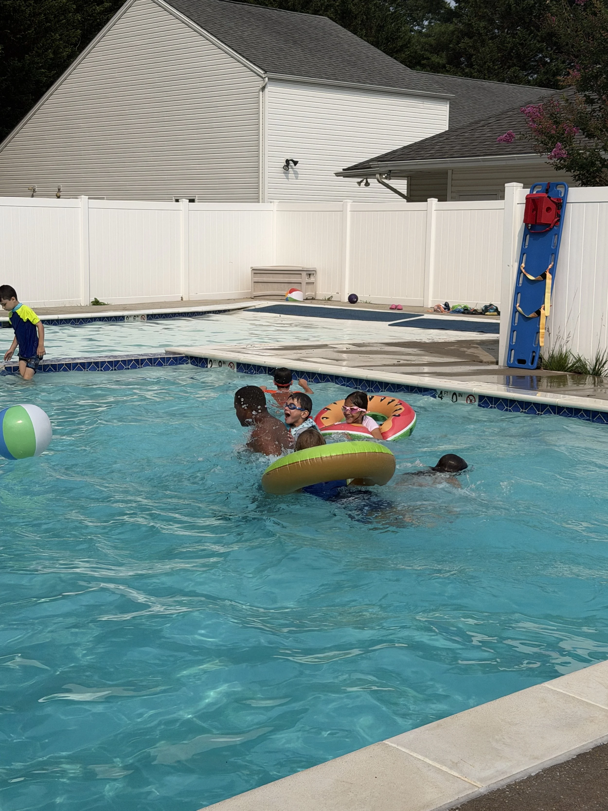Children playing and swimming in a backyard pool with a white fence, pool floaties, and toys visible on the pool deck.