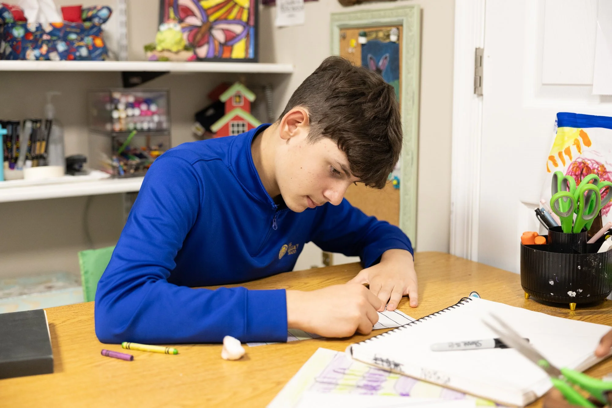 A young boy with brown hair, wearing a blue sweatshirt, is sitting at a wooden table and drawing or writing in a spiral notebook with a black pen. The background features art supplies on shelves and colorful artwork on the walls, including a butterfly painting.