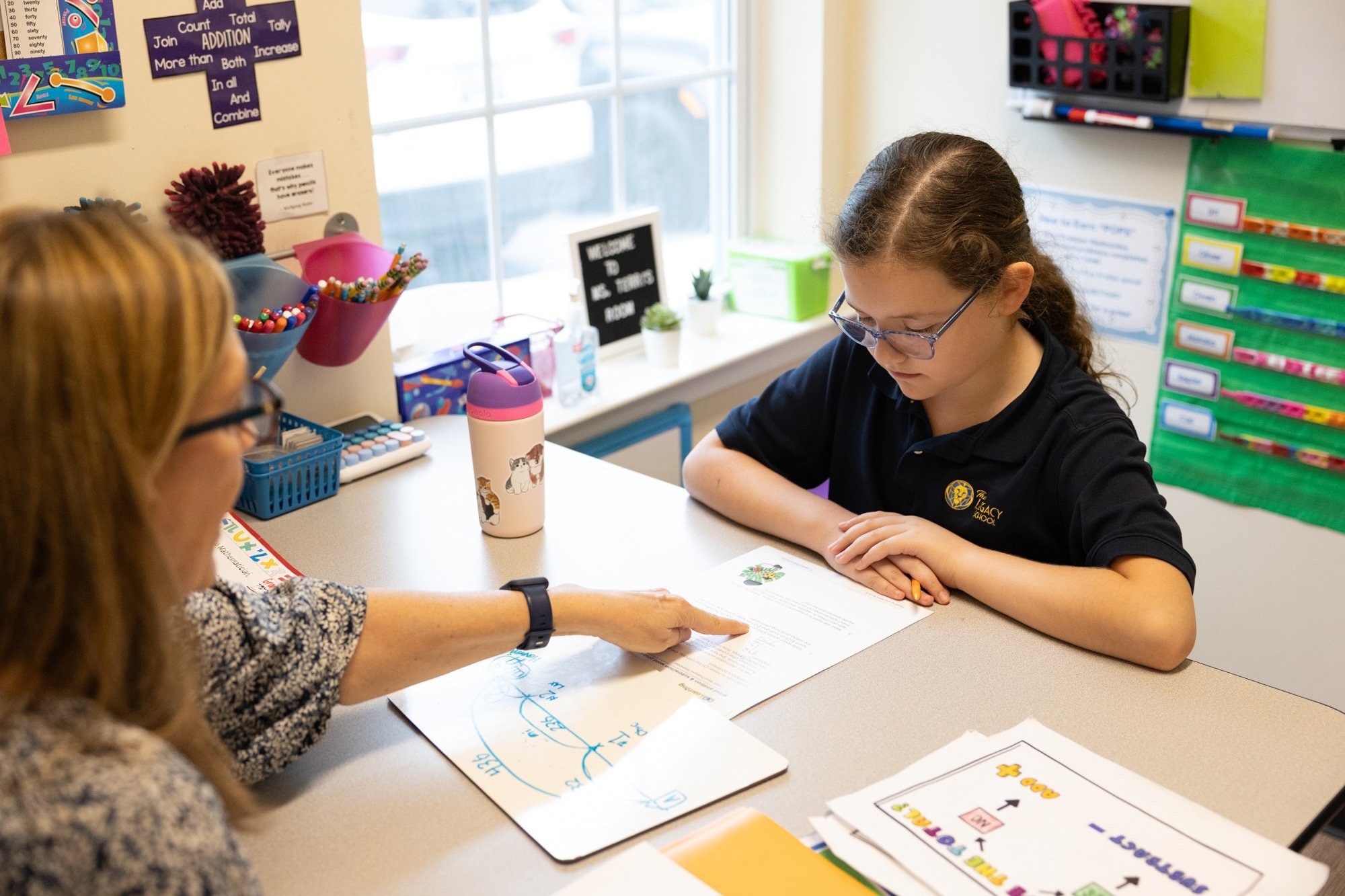 A young girl with glasses and a dark polo shirt sits at a classroom table, looking at a worksheet with colorful drawings and text. An adult woman with glasses and a wristwatch points at the worksheet, engaging with the girl. On the table are various school supplies, including colored markers, a pink and white water bottle with animal illustrations, and other educational materials. The classroom has bright natural light from a window and colorful educational posters and supplies on the walls.