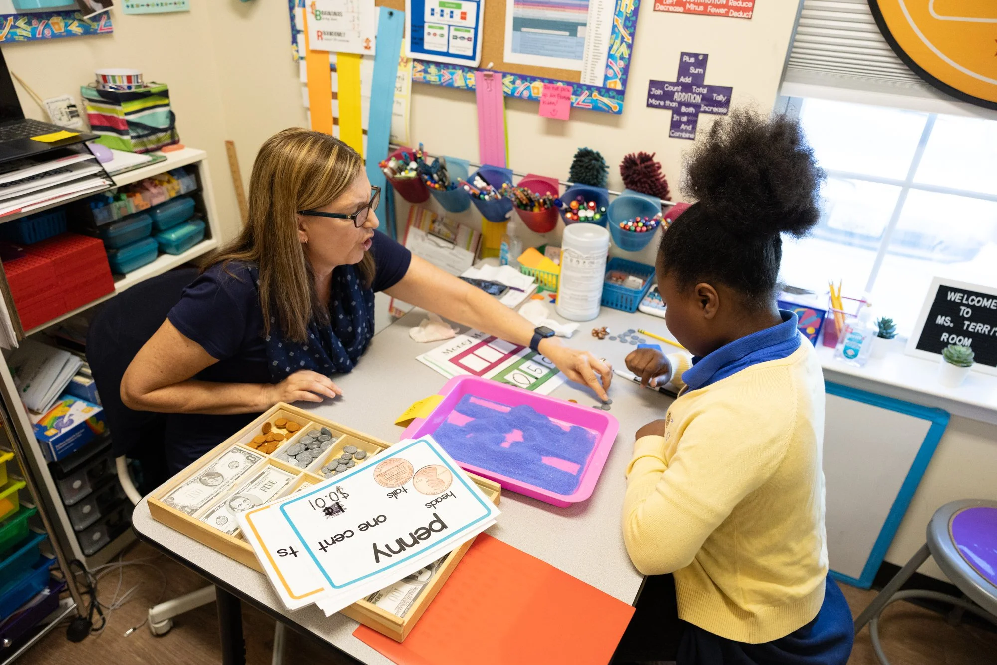 A classroom scene with a teacher and a student engaging in a learning activity at a table. The table has various educational materials, including play money and signs related to money, with a colorful bulletin board and supplies in the background.