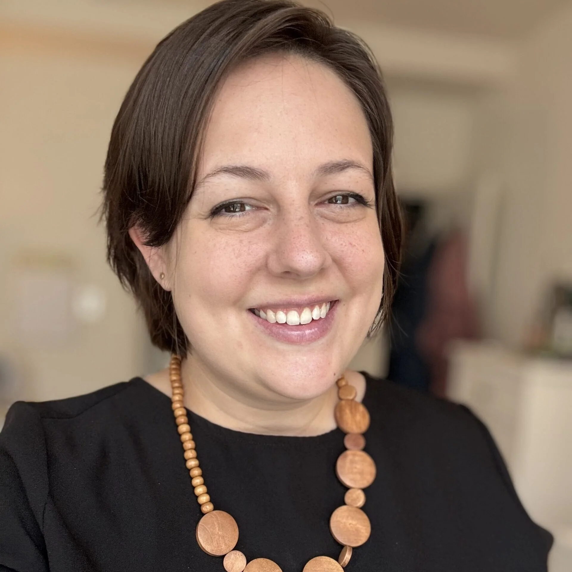 Close-up of a smiling woman with short brown hair wearing a black top and wooden bead jewelry.